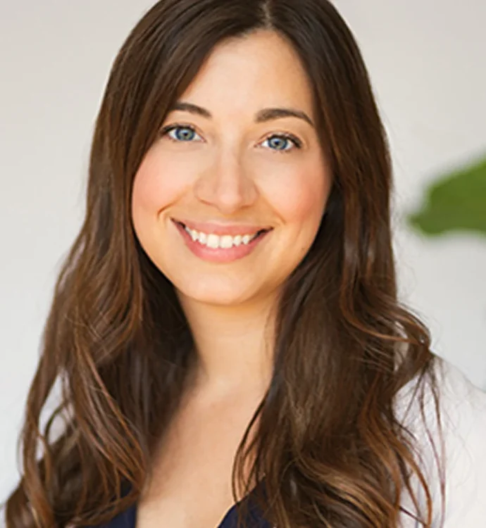 Smiling woman with long brown hair, wearing a blue top, against a light background.