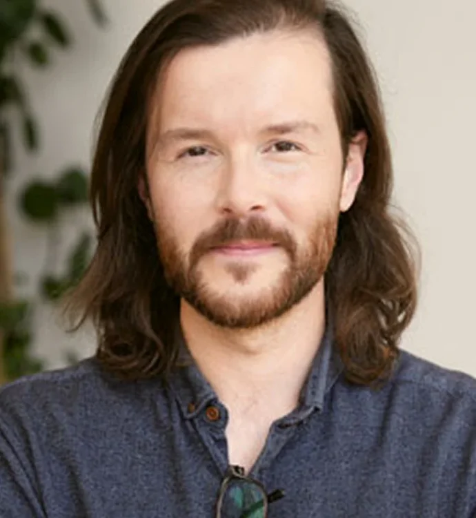 Portrait of a bearded man with long hair and a calm expression, wearing a blue shirt.