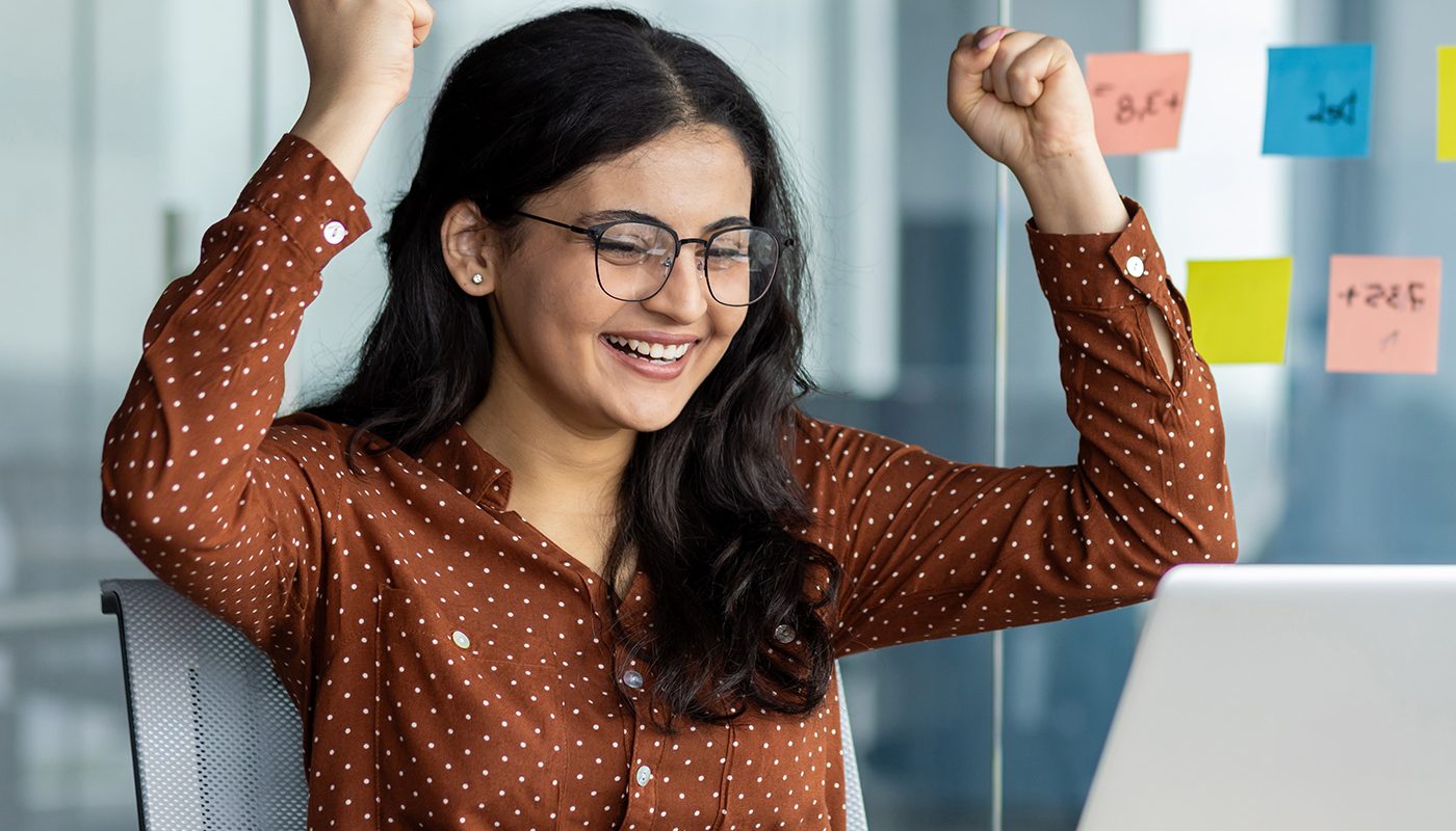 Excited woman celebrates success at laptop in modern office with sticky notes on glass wall.