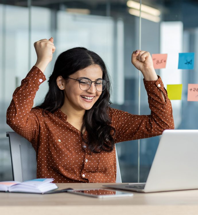 Excited woman celebrates success at laptop in modern office with sticky notes on glass wall.