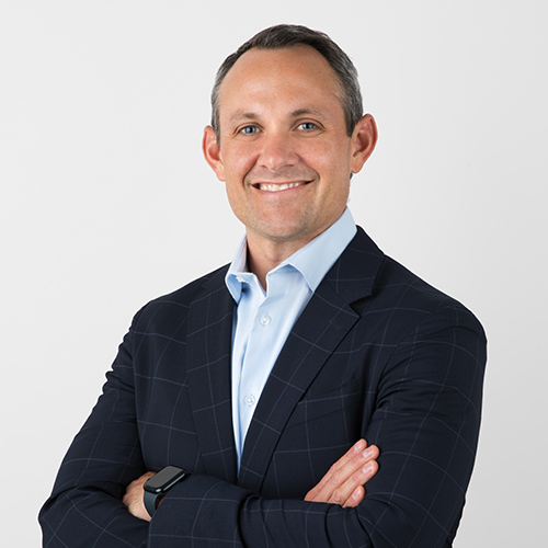 Man in a suit smiling with arms crossed against a plain background.
