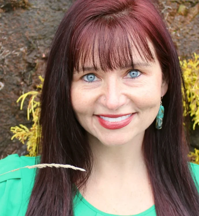 Smiling woman with red hair, green top, and teal earrings, standing outdoors by a wall with plants.