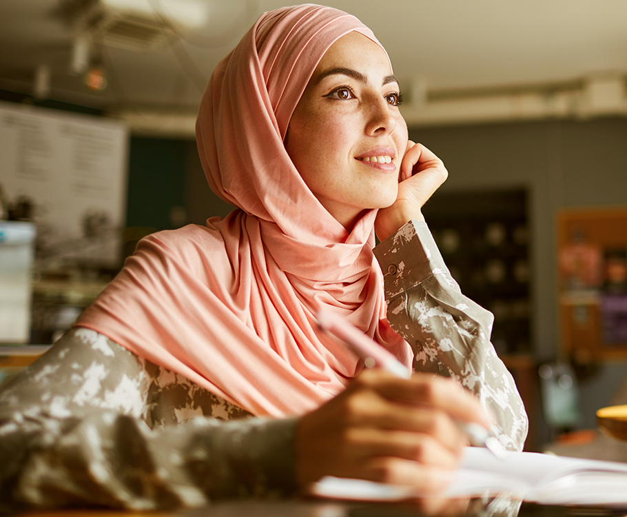 Woman in a pink hijab smiling and writing, looking inspired in a cozy indoor setting.