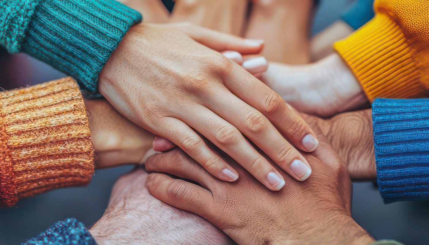 Hands of diverse people stacked in unity, wearing colorful sweaters.