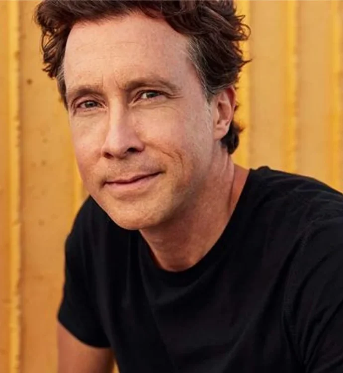 Man in a black t-shirt smiling in front of an orange wall.