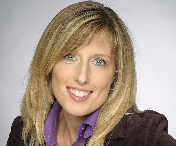 Smiling woman with blonde hair, wearing a purple shirt and brown jacket, neutral background.