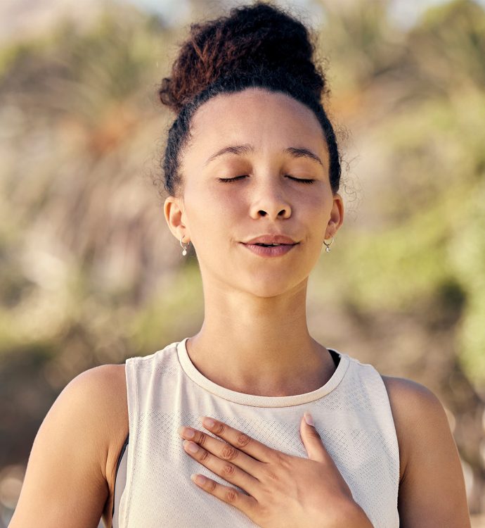 Woman meditating outdoors with eyes closed, hand on chest, and peaceful expression.