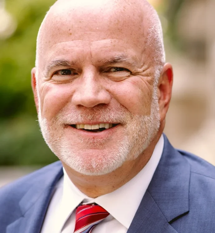 Bald man in a suit smiling outdoors with a background of greenery.