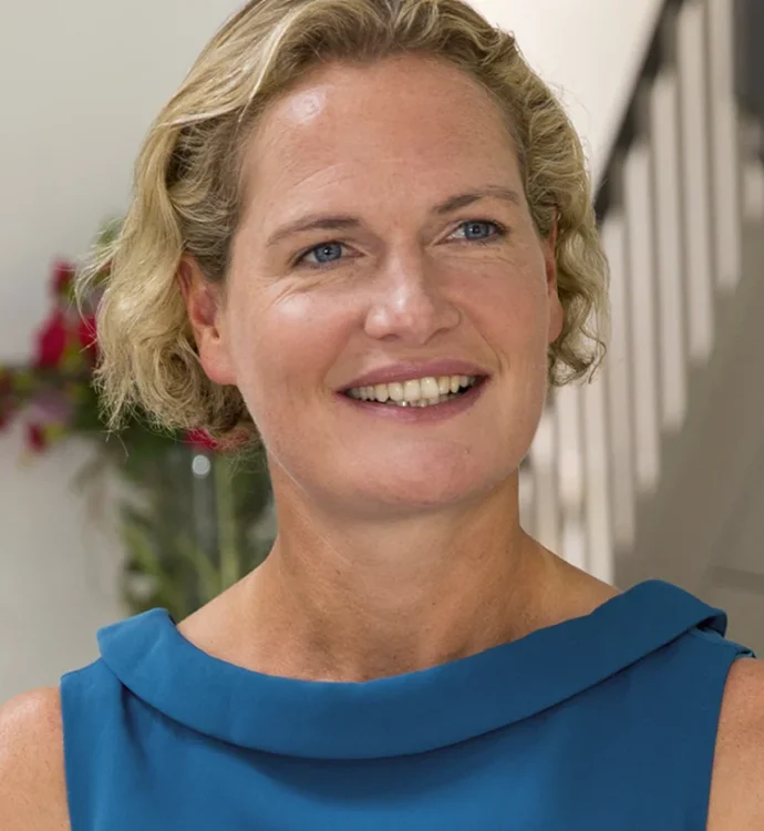 Smiling woman in blue top standing indoors with flowers in background.