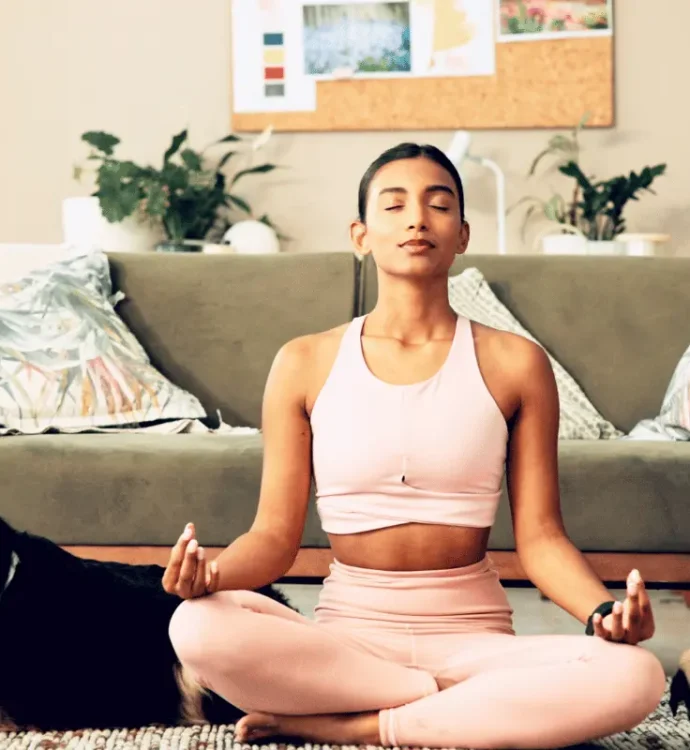 A woman sits cross-legged in a relaxed meditation pose on a rug in front of a couch, eyes closed, with two dogs nearby. The setting is a cozy, plant-filled living room, conveying calm and groundedness.