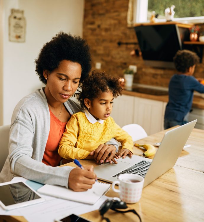 Mom working at home with kids; multitasking with laptop, notebook, and child on lap.