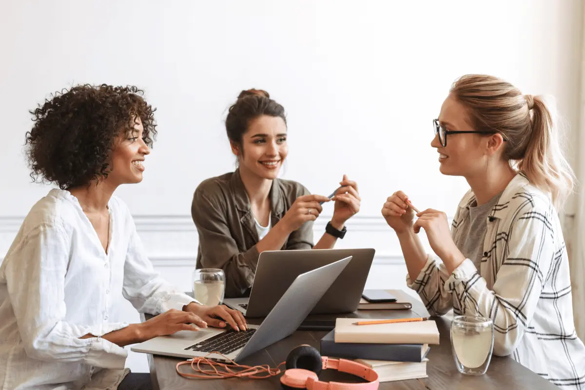 Three women smiling and discussing work around laptops in a meeting.