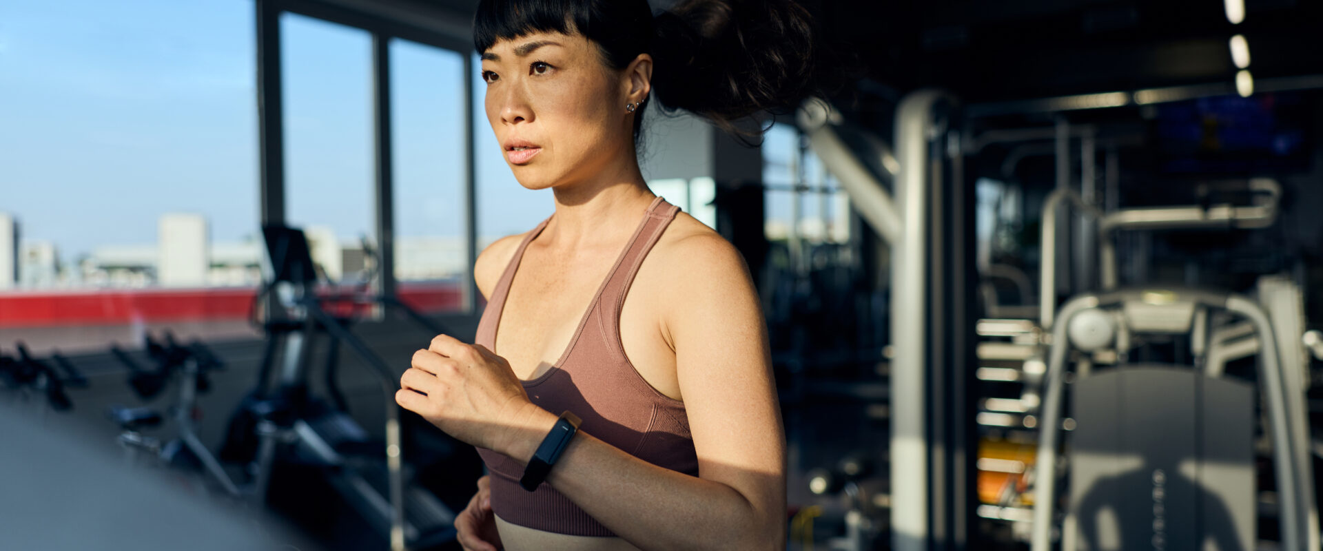 Woman running on treadmill in gym with city view through windows.