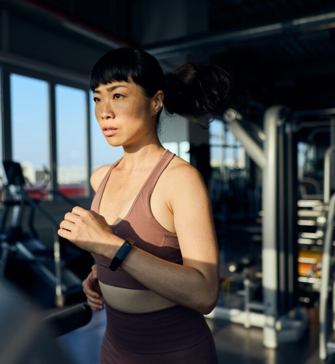 Woman running on treadmill in gym with city view through windows.
