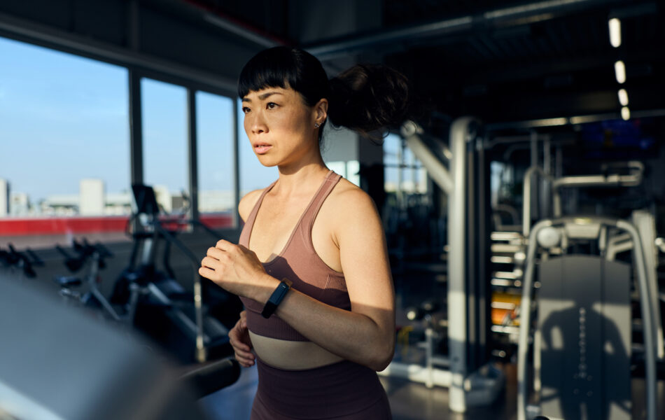 Woman running on treadmill in gym with city view through windows.