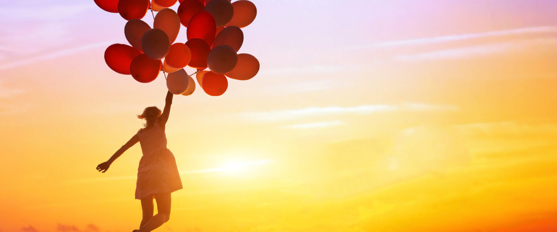 Person holding balloons on a beach during a colorful sunset.