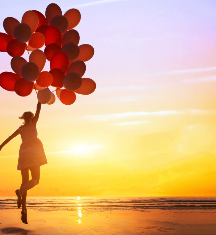 Person holding balloons on a beach during a colorful sunset.