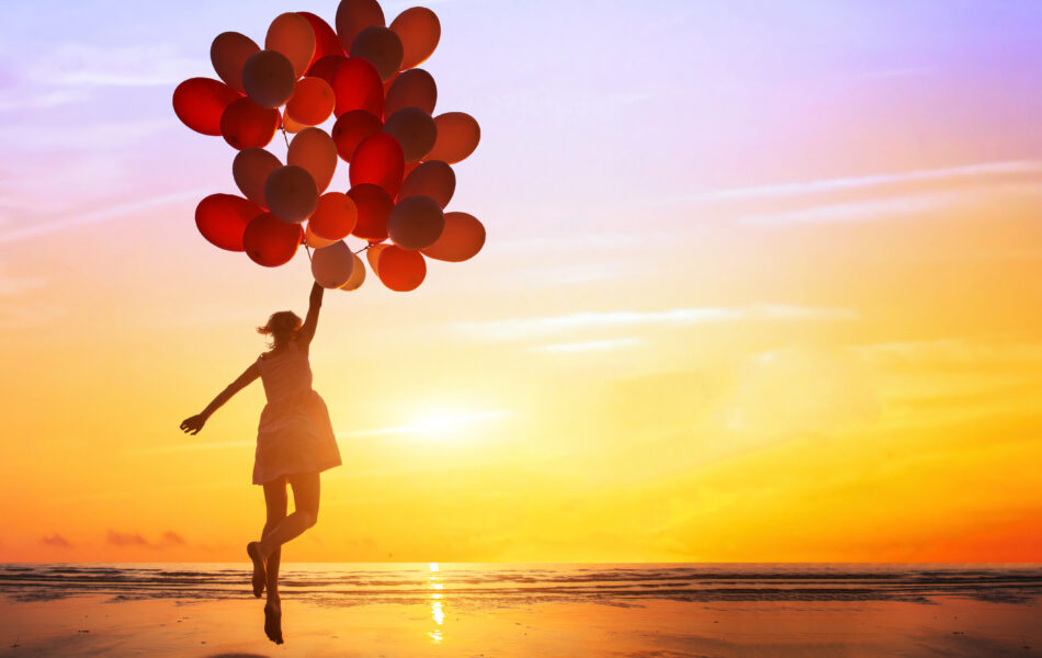 Person holding balloons on a beach during a colorful sunset.