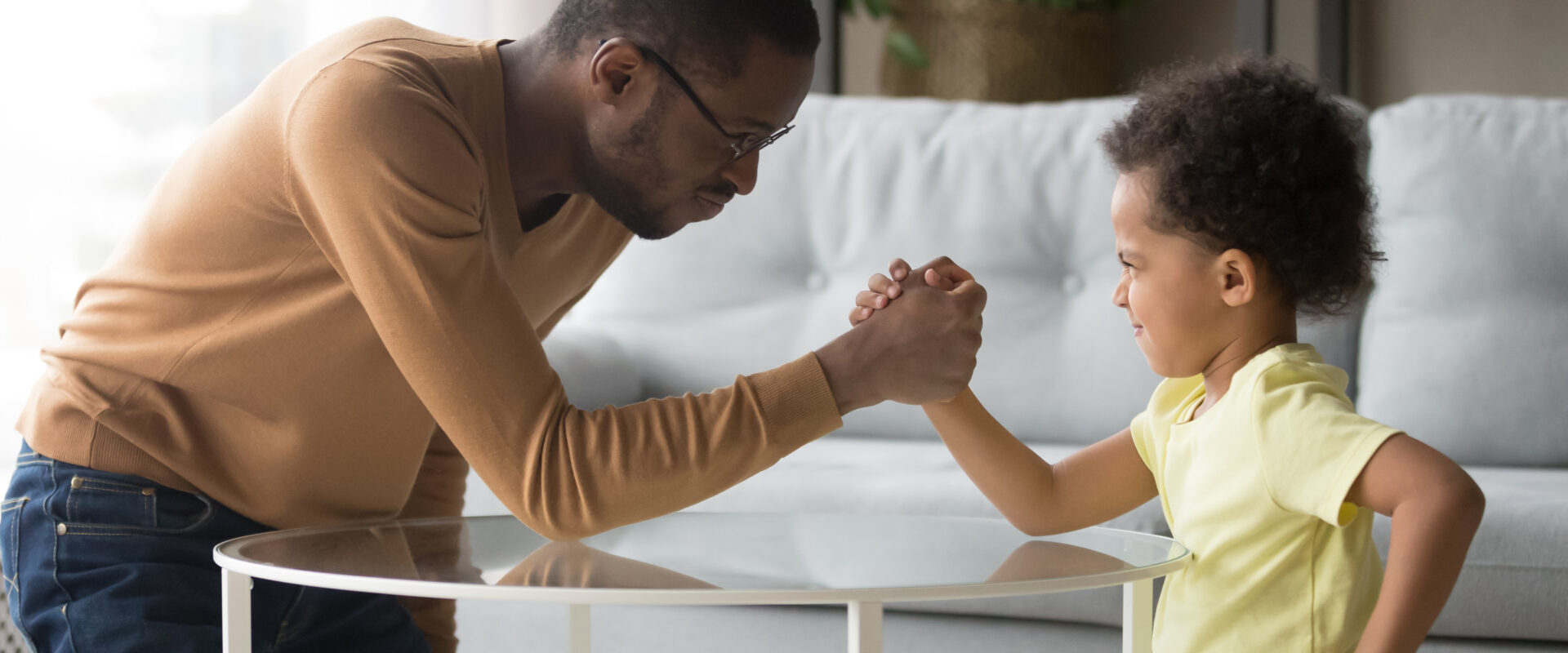 Father and child arm wrestling in living room, smiling and playful.