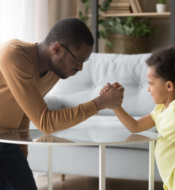 Father and child arm wrestling in living room, smiling and playful.
