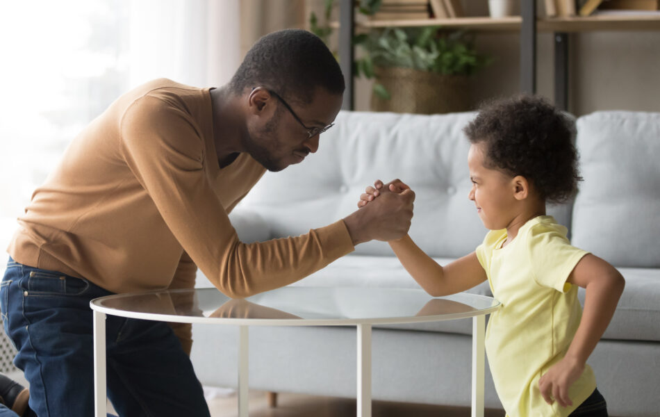 Father and child arm wrestling in living room, smiling and playful.
