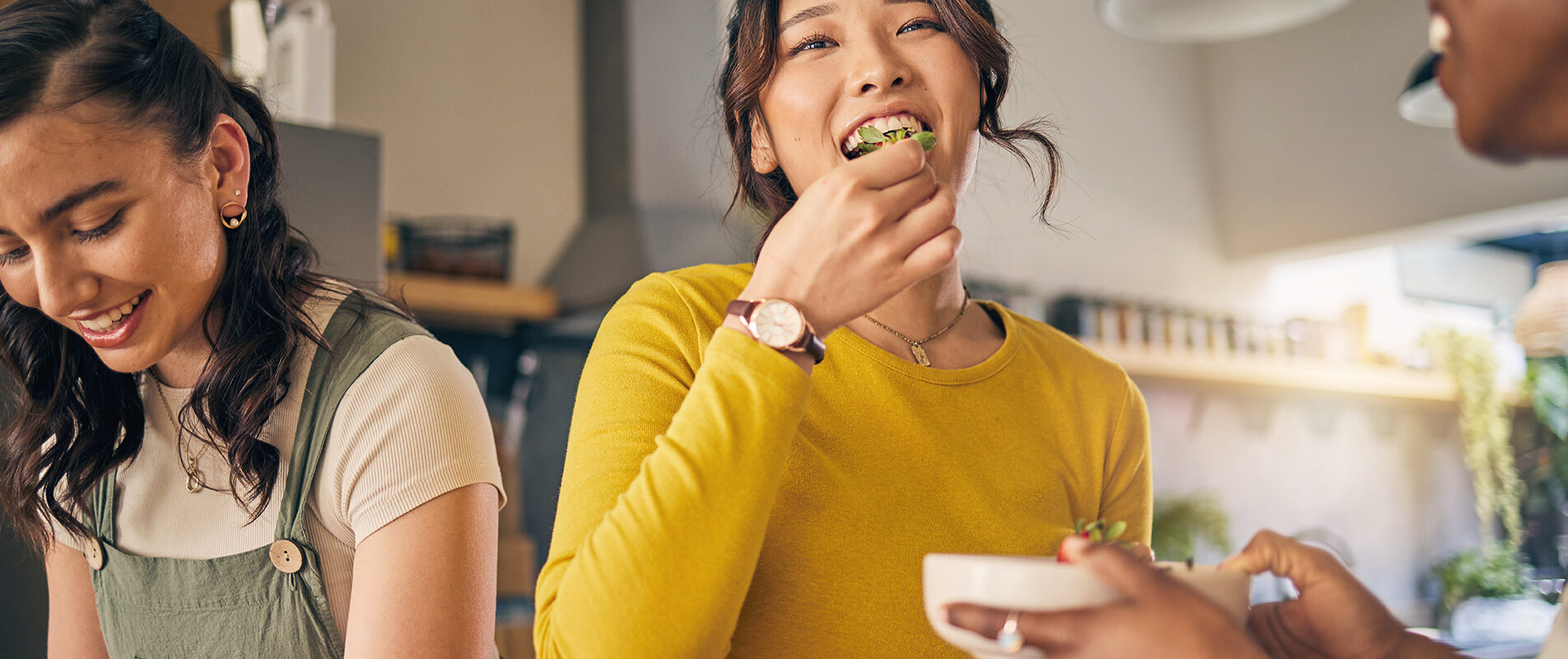 Three young women are in a bright kitchen preparing and eating food together. One woman in a yellow top smiles while eating a piece of salad, another on the left looks down while working with ingredients, and a third (partially visible) holds a bowl. The setting feels casual and friendly, with warm lighting and kitchen shelves in the background.