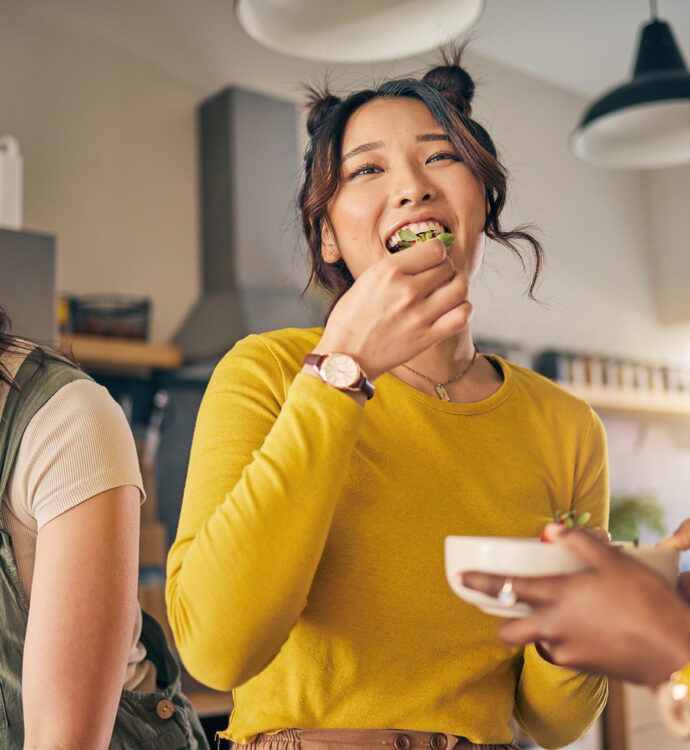 Three young women are in a bright kitchen preparing and eating food together. One woman in a yellow top smiles while eating a piece of salad, another on the left looks down while working with ingredients, and a third (partially visible) holds a bowl. The setting feels casual and friendly, with warm lighting and kitchen shelves in the background.