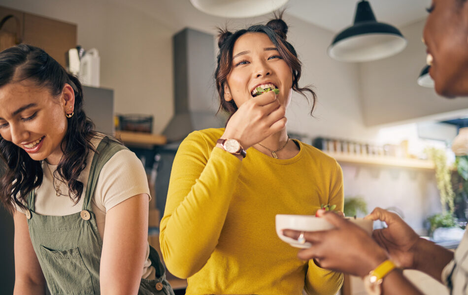 Three young women are in a bright kitchen preparing and eating food together. One woman in a yellow top smiles while eating a piece of salad, another on the left looks down while working with ingredients, and a third (partially visible) holds a bowl. The setting feels casual and friendly, with warm lighting and kitchen shelves in the background.