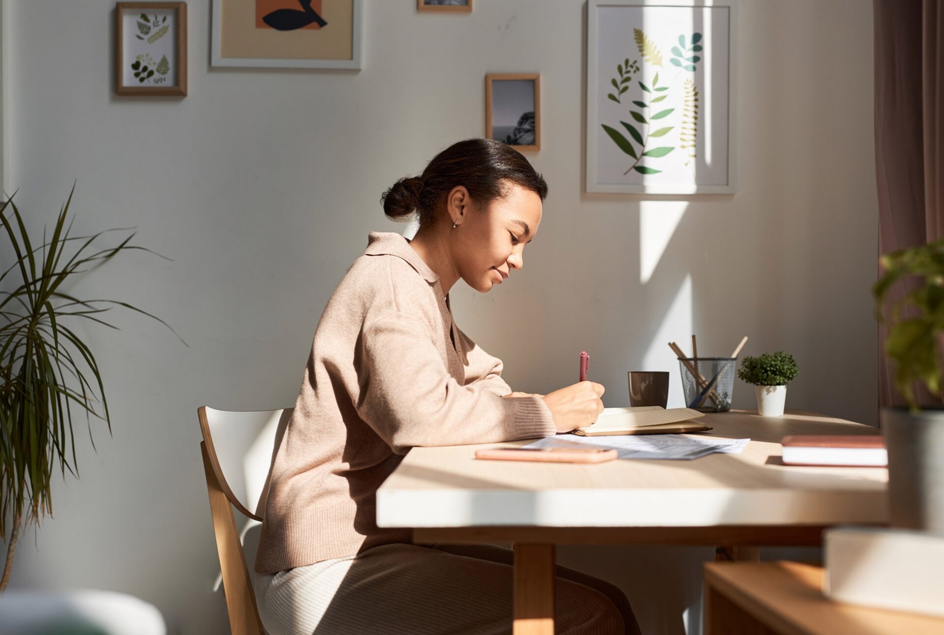 Woman writing at sunlit desk with plants and framed pictures.