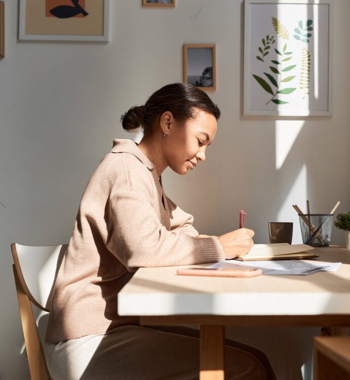 Woman writing at sunlit desk with plants and framed pictures.
