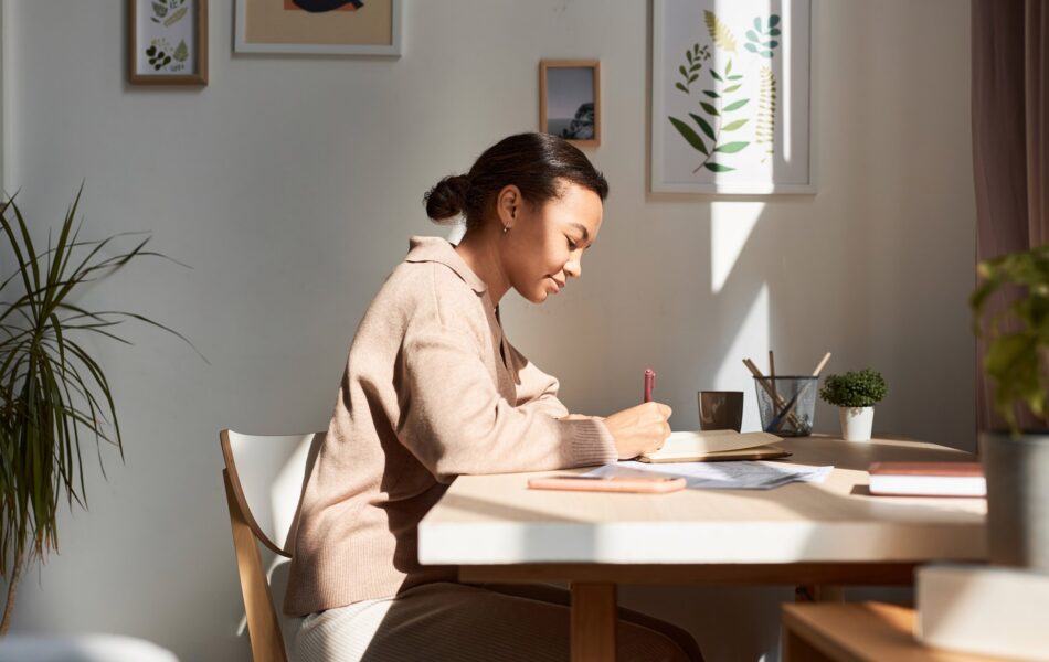 Woman writing at sunlit desk with plants and framed pictures.