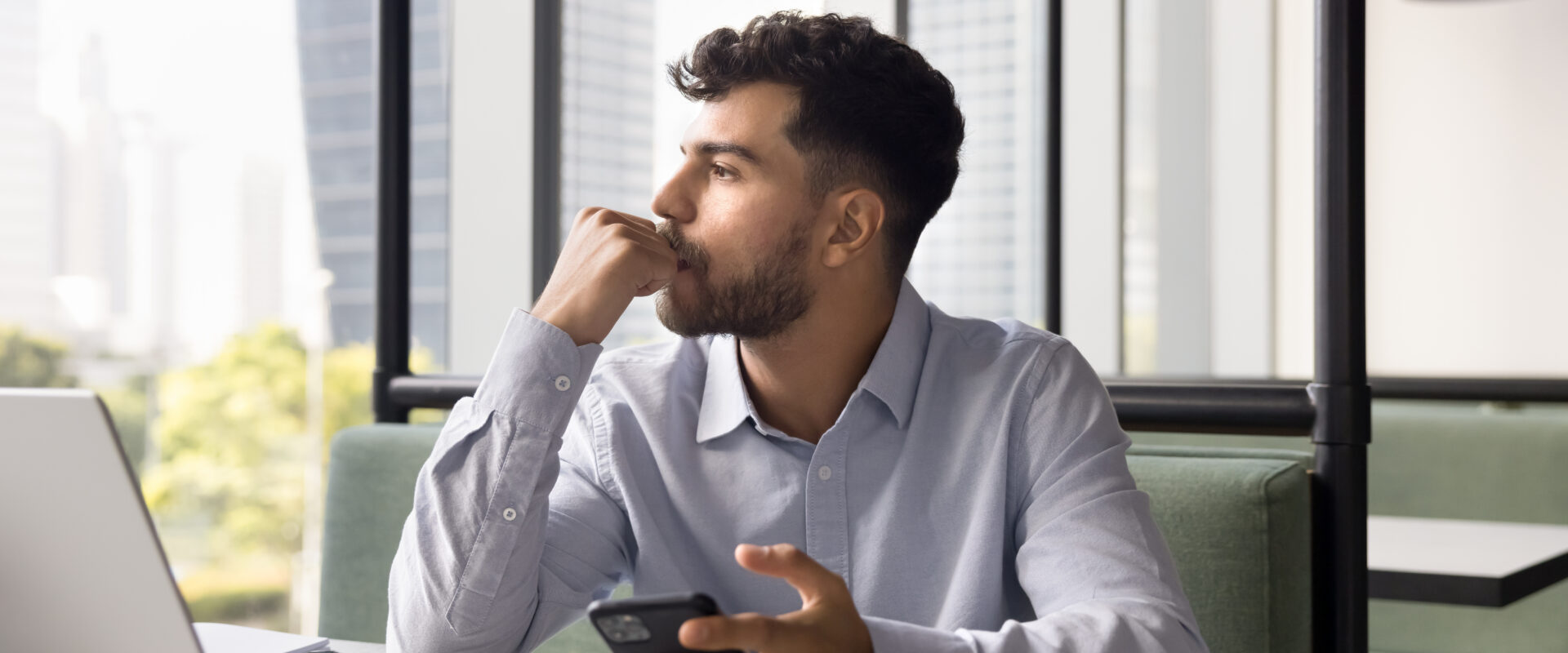 Man contemplating while holding phone at a desk with a laptop in an office setting.