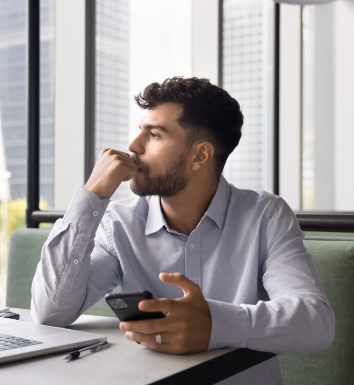 Man contemplating while holding phone at a desk with a laptop in an office setting.