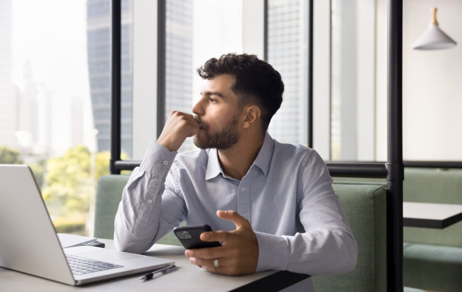 Man contemplating while holding phone at a desk with a laptop in an office setting.