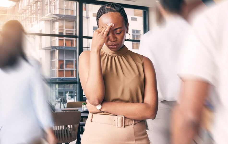 A woman stands indoors with her hand pressed to her forehead and eyes closed, appearing stressed or overwhelmed, while blurred people move around her in the foreground. Large windows and apartment buildings are visible in the background.