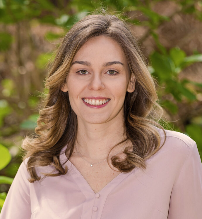 Smiling woman outdoors with curly hair and pink blouse, in front of greenery and soft background.
