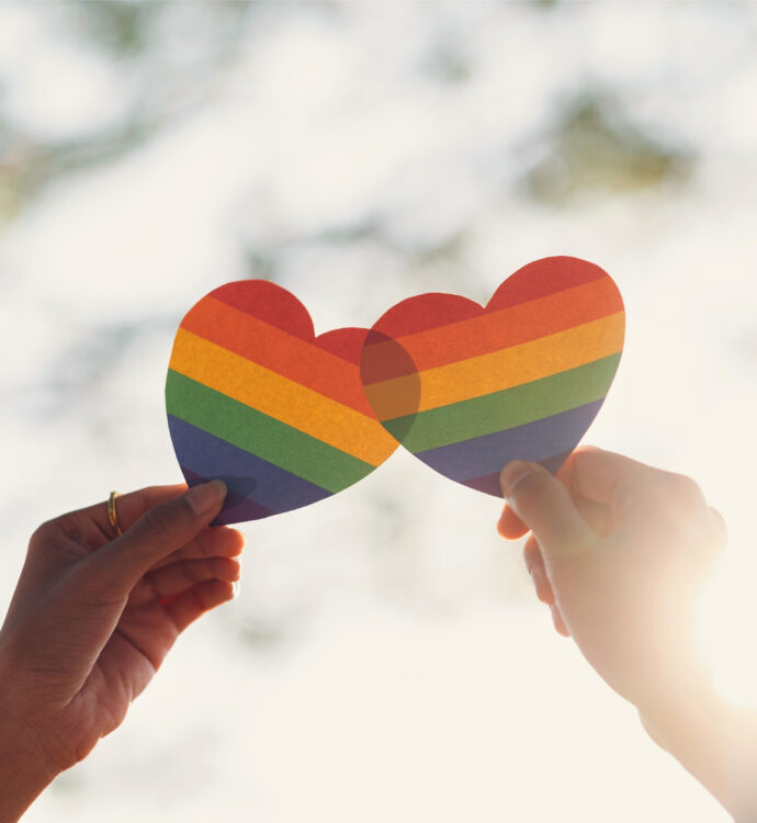 Two hands holding rainbow heart cutouts in sunlight, symbolizing LGBTQ+ pride and love.