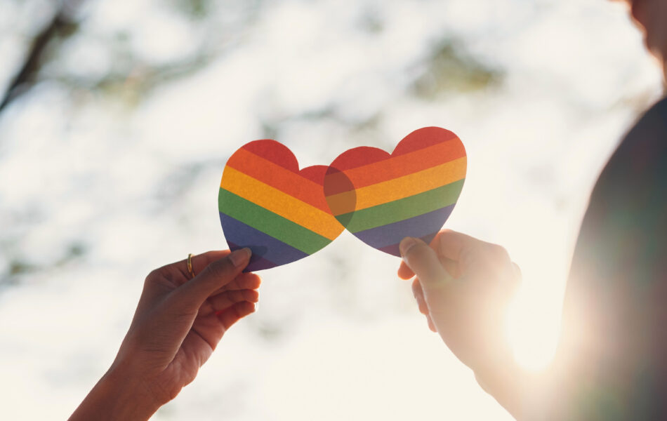 Two hands holding rainbow heart cutouts in sunlight, symbolizing LGBTQ+ pride and love.