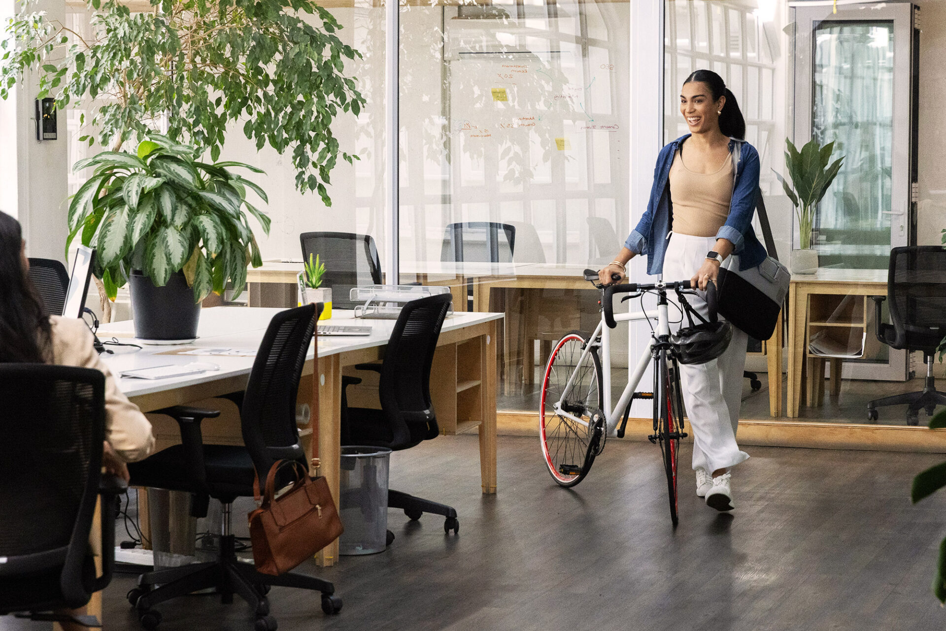 Employee walking her bicycle into a plant-filled open office, smiling as she greets a colleague at a shared desk