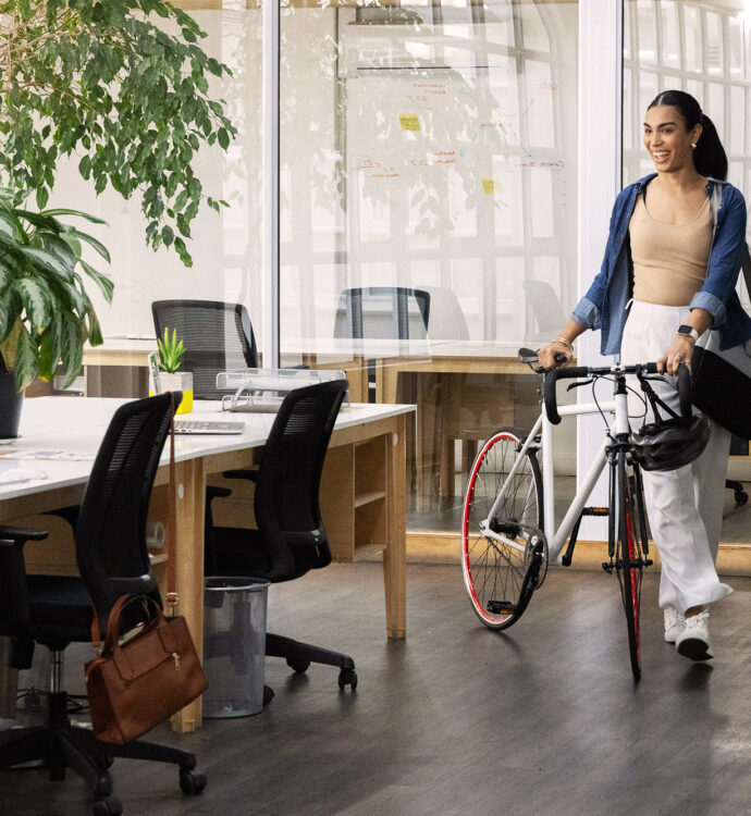 Employee walking her bicycle into a plant-filled open office, smiling as she greets a colleague at a shared desk