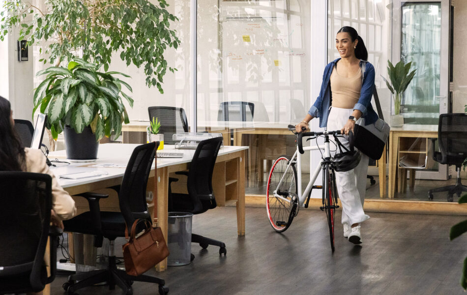 Employee walking her bicycle into a plant-filled open office, smiling as she greets a colleague at a shared desk
