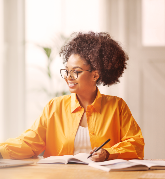 Woman in a yellow shirt smiling at her laptop while taking notes at a desk.