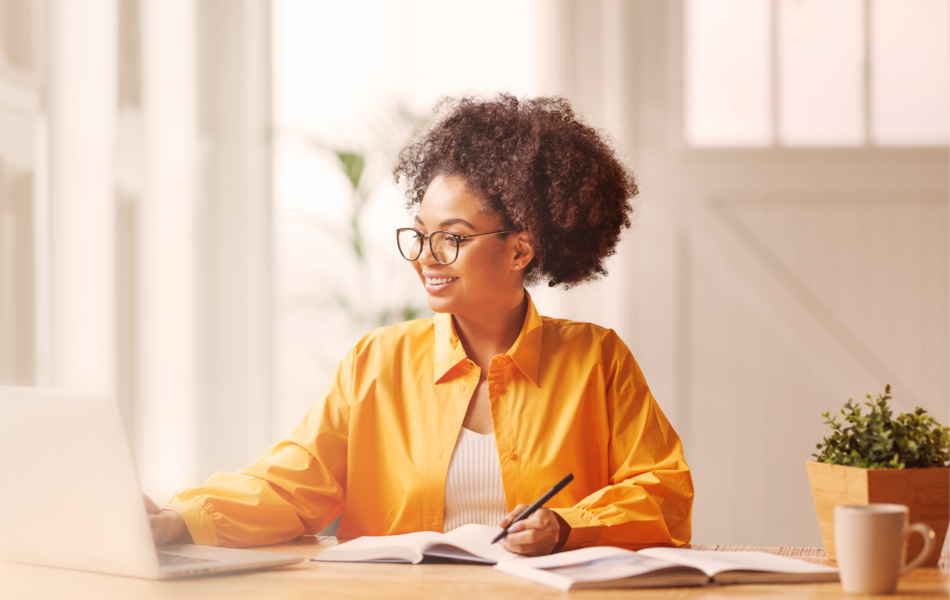 Woman in a yellow shirt smiling at her laptop while taking notes at a desk.