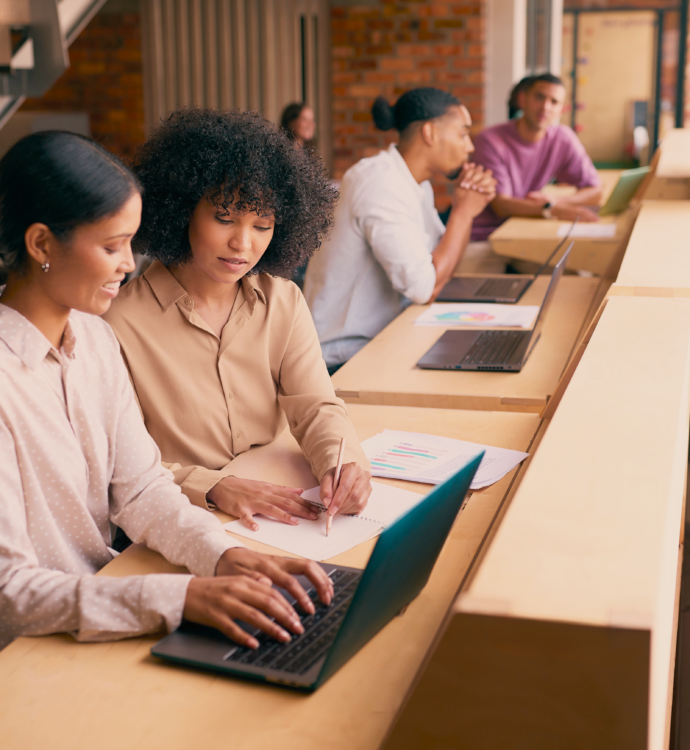 Two colleagues reviewing notes and a laptop together at a shared desk, with coworkers working nearby in an open office.