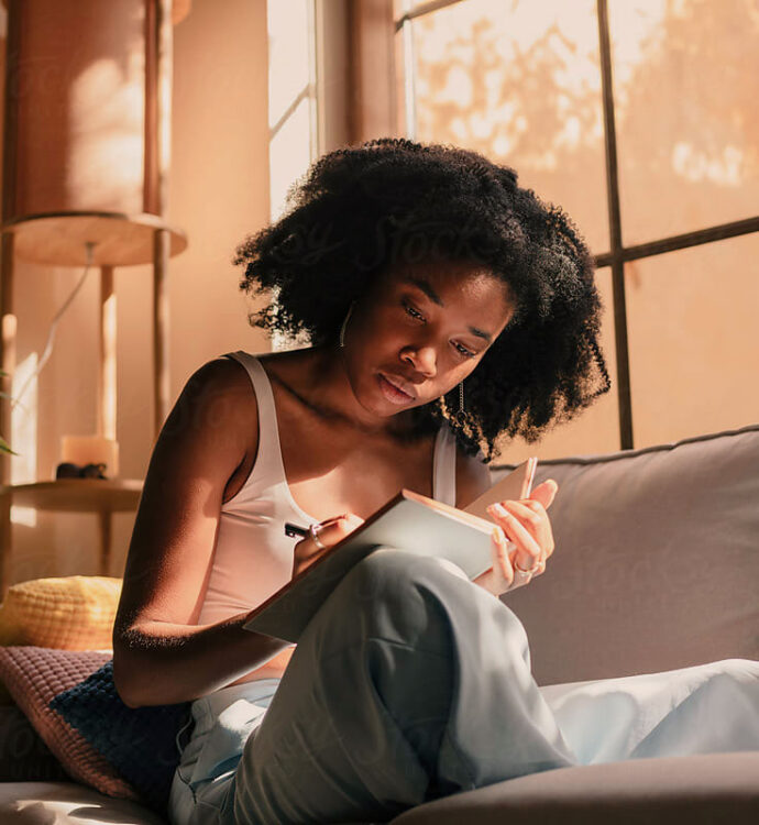 Woman sitting on her couch at home, writing in a notebook in warm afternoon light.