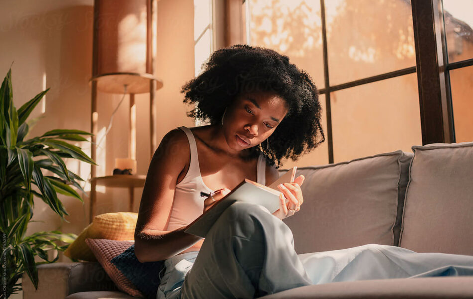Woman sitting on her couch at home, writing in a notebook in warm afternoon light.