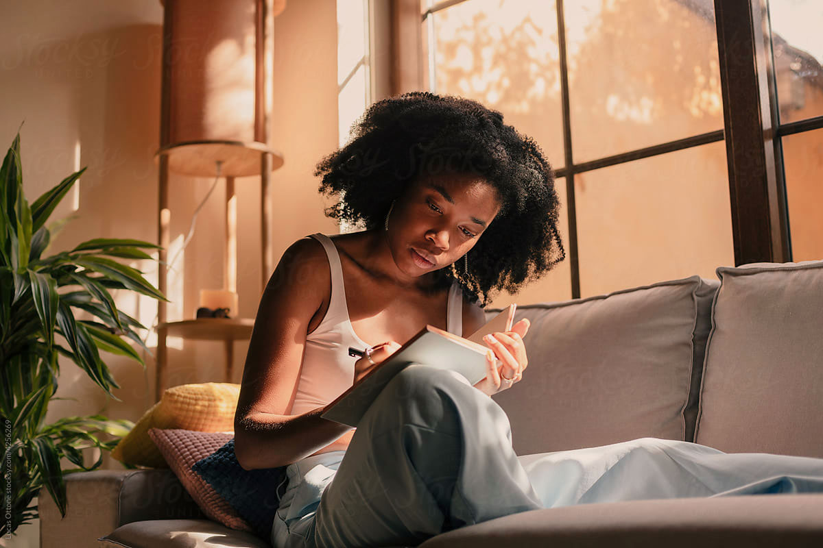 Woman sitting on her couch at home, writing in a notebook in warm afternoon light.