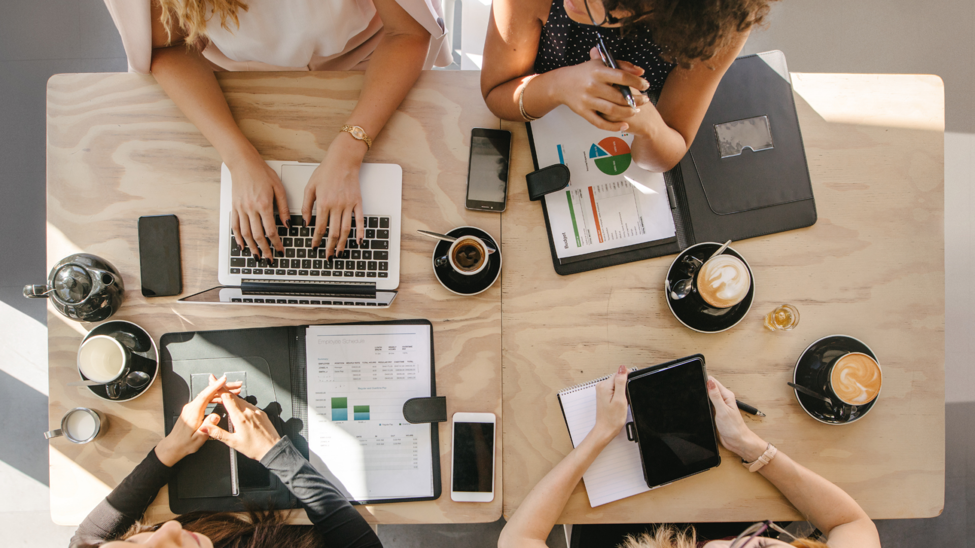 Overhead view of coworkers at a table reviewing budget and scheduling documents with laptops and coffee.