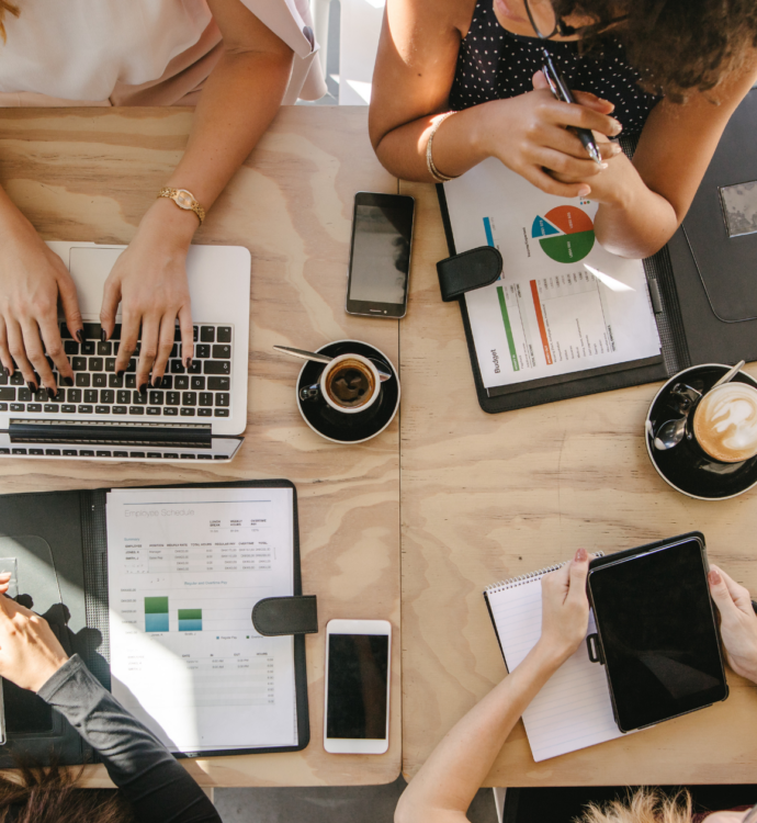 Overhead view of coworkers at a table reviewing budget and scheduling documents with laptops and coffee.