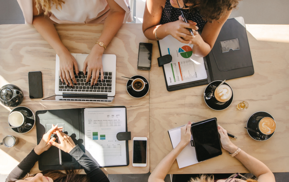 Overhead view of coworkers at a table reviewing budget and scheduling documents with laptops and coffee.